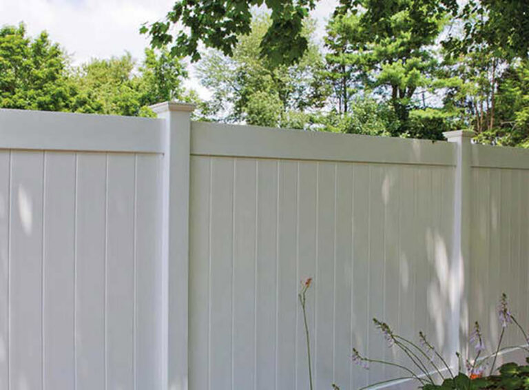 White vinyl privacy fence with trees overhead and flowering plants in front.