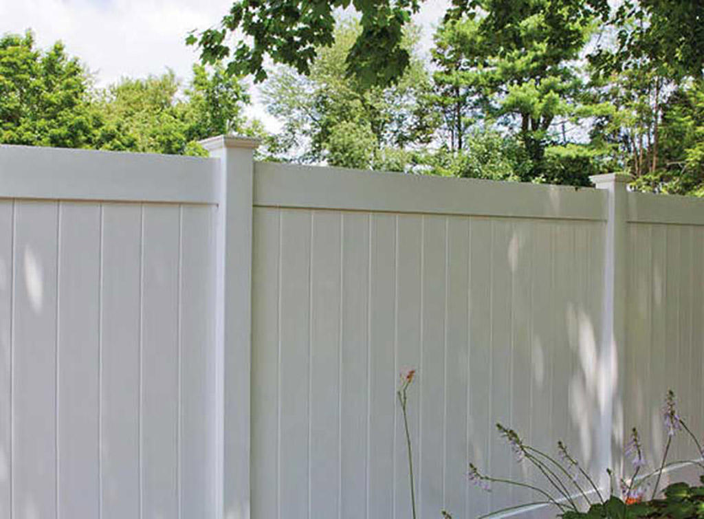 White vinyl privacy fence with trees overhead and flowering plants in front.